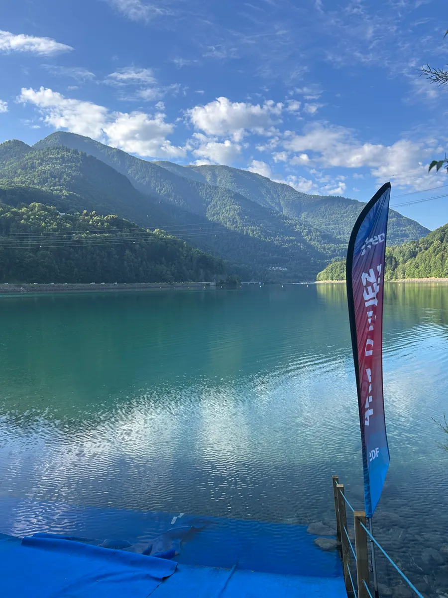 Blick über die Talsperre mit Bergen im Hintergrund, im Vordergrund eine Fahne am Steg